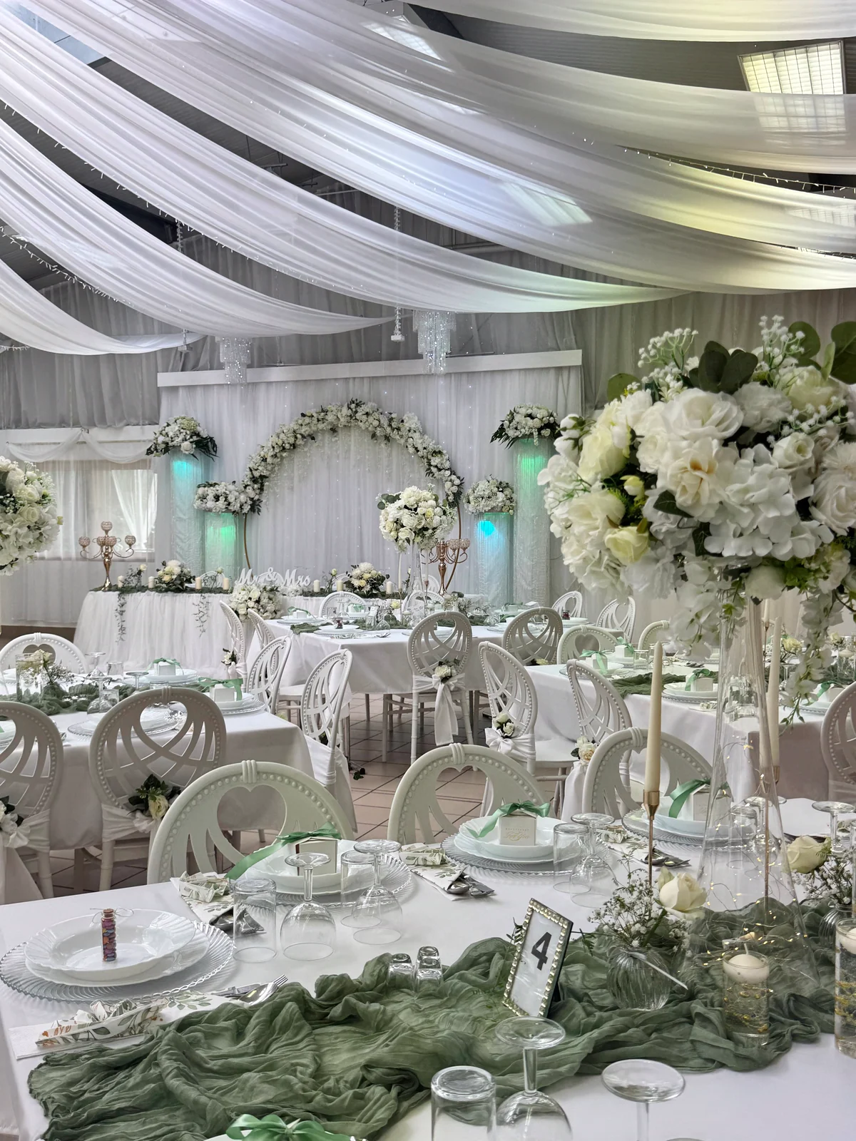 Festlich gedeckte Tische mit weißen Blumengestecken und salbeigrünen Tischläufern, im Hintergrund der Blumenbogen