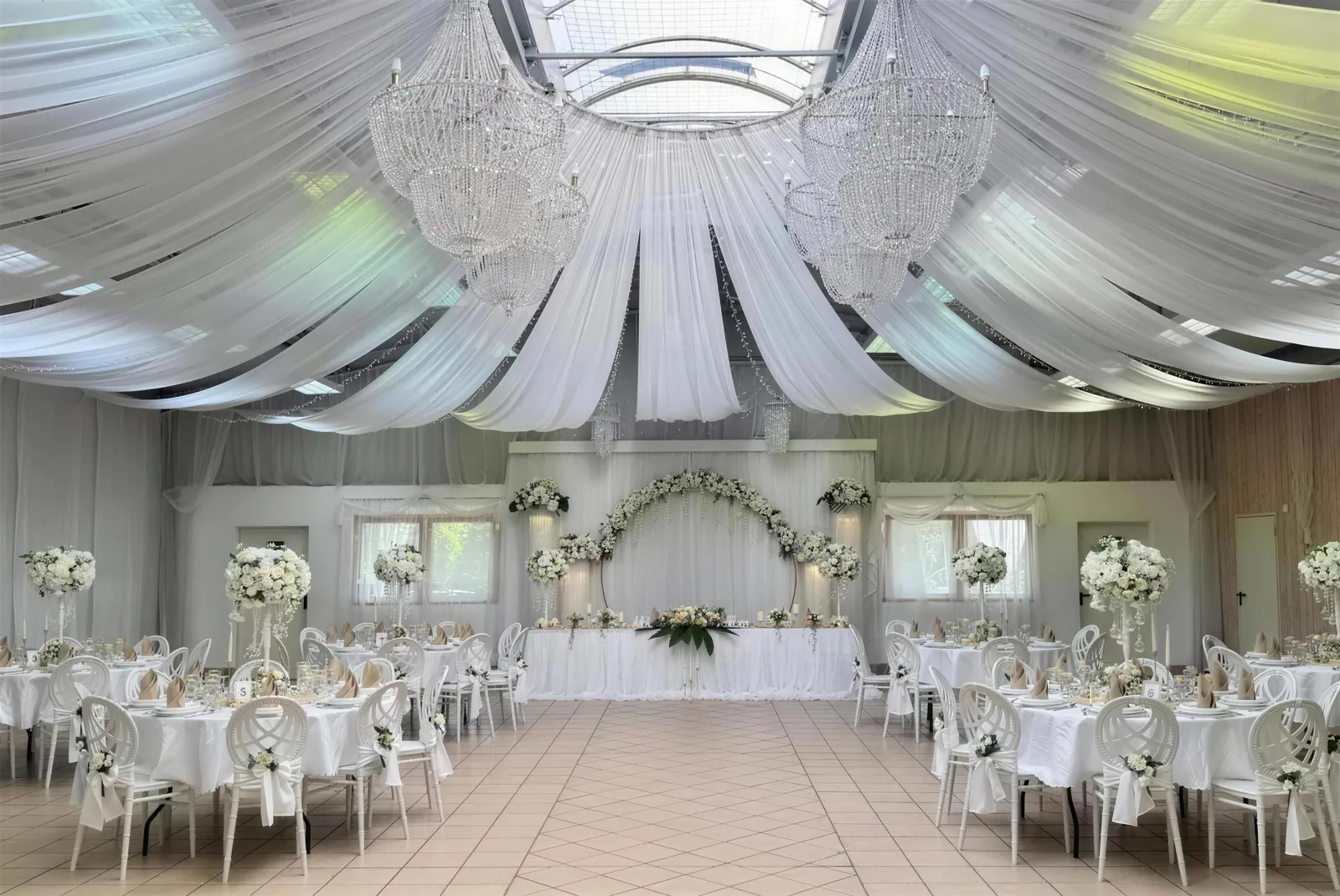 Die Eventhalle im Hochzeits-Setup mit Kronleuchtern, weißer Stoffdecke und Blumenbogen